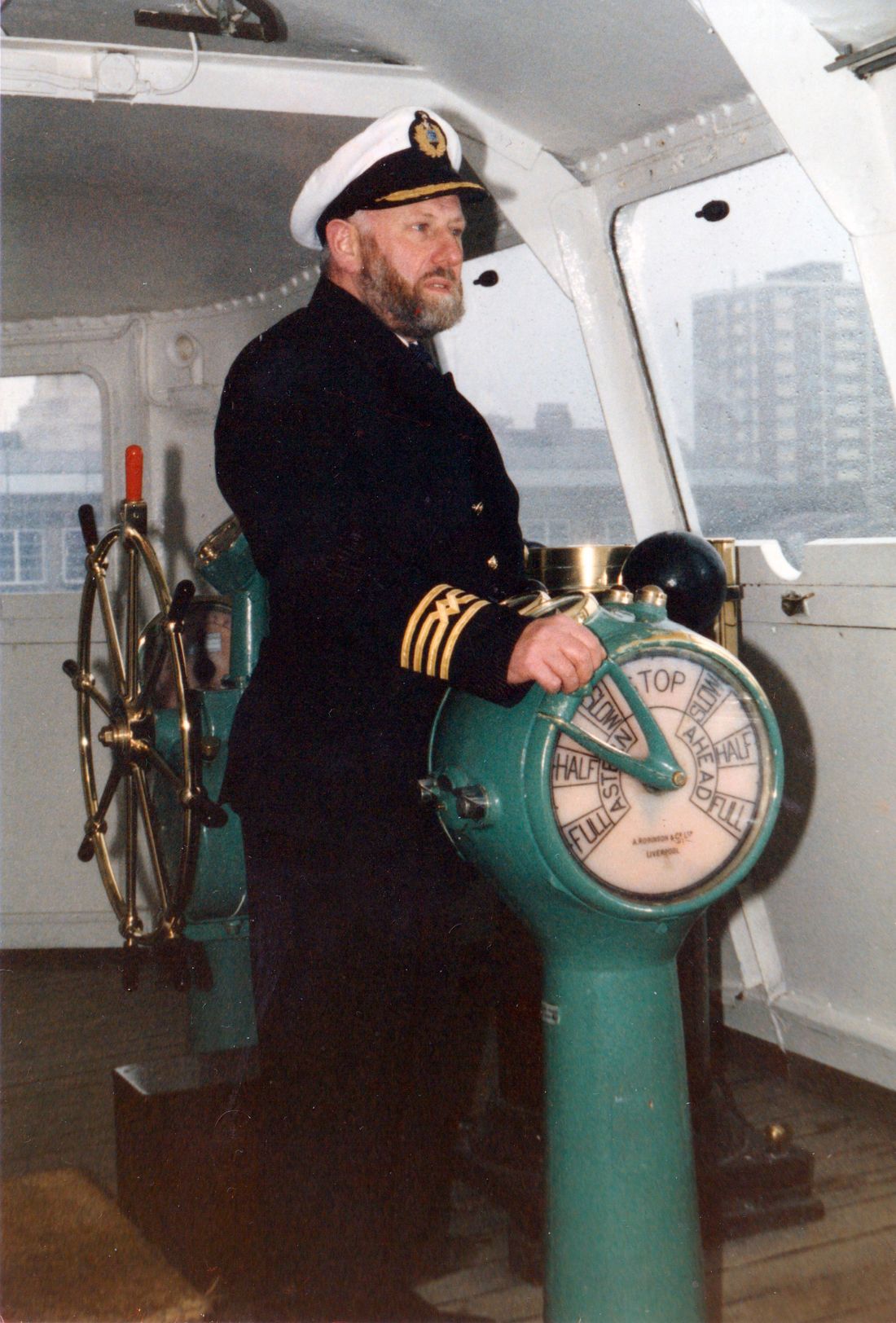 Captain Mike Lowe in uniform and cap, bearded, standing in the wheelhouse of the MV Royal Iris with his hand on the green engine-room telegraph and the ship's wheel behind him.