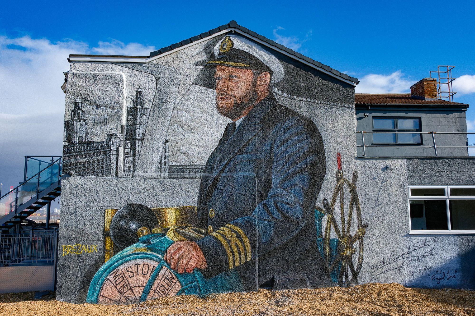 The Brezaux mural of Captain Mike Lowe on the gable end of the Master Mariner, under a blue sky, with the Liver Building behind him and the telegraph at the bottom left.