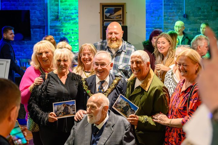 Mike Lowe seated in front of the Mayor of Wirral, family and friends, two of them holding up the postcard of the mural.